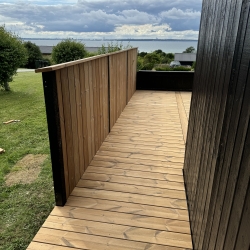 Stor træterrasse mellem sort hus med arbejdsbil i baggrunden.