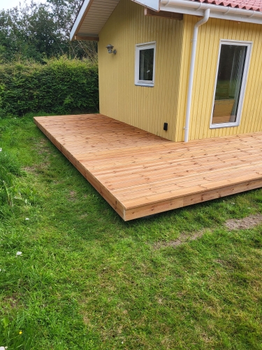 Lang træterrasse ved gult hus med flere havedøre