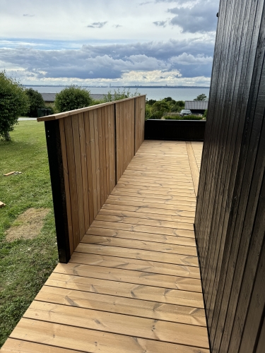 Stor træterrasse mellem sort hus med arbejdsbil i baggrunden.